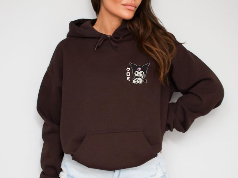 Puede incluir: Una sudadera con capucha marr&oacute;n recortada con un gr&aacute;fico en blanco y negro de un personaje de dibujos animados con el texto japon&eacute;s "クロミ" en la parte delantera.