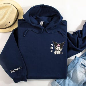 Puede incluir: Una sudadera con capucha azul marino con un personaje de dibujos animados en blanco y negro con mejillas rosadas y el texto japon&eacute;s "Kuromi" en la parte delantera. La sudadera tiene un coraz&oacute;n blanco y la palabra "Name" bordada en la manga izquierda.