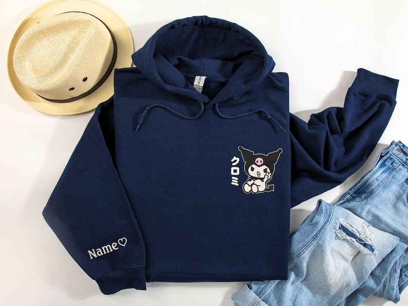 Puede incluir: Una sudadera con capucha azul marino con un personaje de dibujos animados en blanco y negro con mejillas rosadas y el texto japon&eacute;s "Kuromi" en la parte delantera. La sudadera tiene un coraz&oacute;n blanco y la palabra "Name" bordada en la manga izquierda.