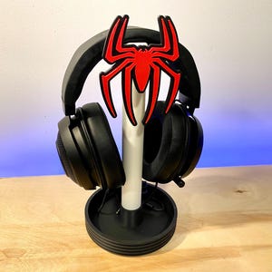 May include: Black headphone stand with a red Spider-Man logo. The stand has a white base and a black ring around the base.