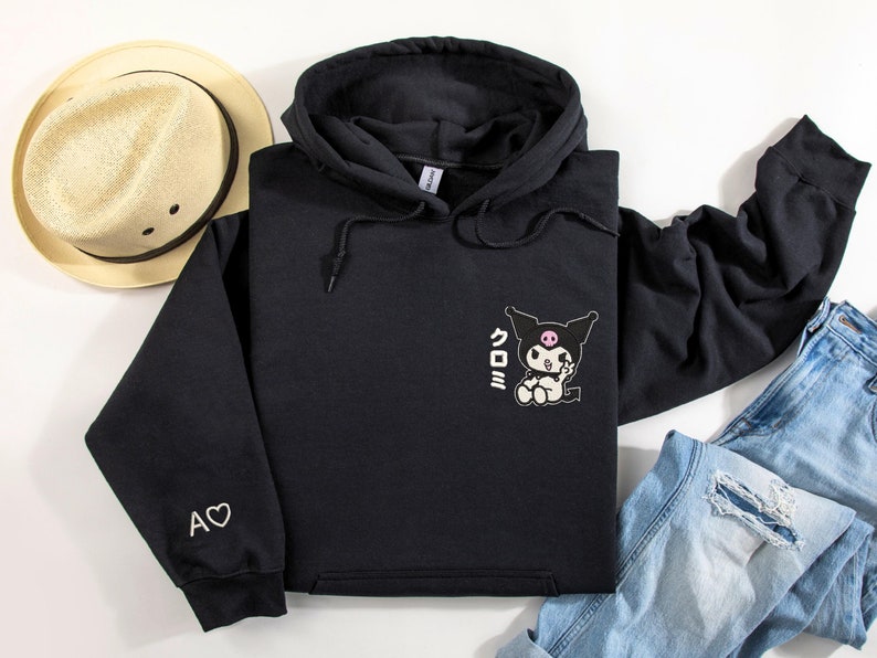 Puede incluir: Sudadera con capucha negra con un coraz&oacute;n blanco y el texto "A" en la manga izquierda. La parte delantera de la sudadera presenta un personaje de dibujos animados en blanco y negro con el texto "クロミ" en japon&eacute;s.