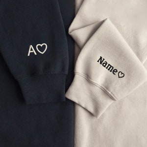 Puede incluir: Dos sudaderas con corazones y texto bordados. La sudadera de la izquierda es azul marino con bordado blanco que dice "A" con un coraz&oacute;n. La sudadera de la derecha es beige claro con bordado negro que dice "Name" con un coraz&oacute;n.