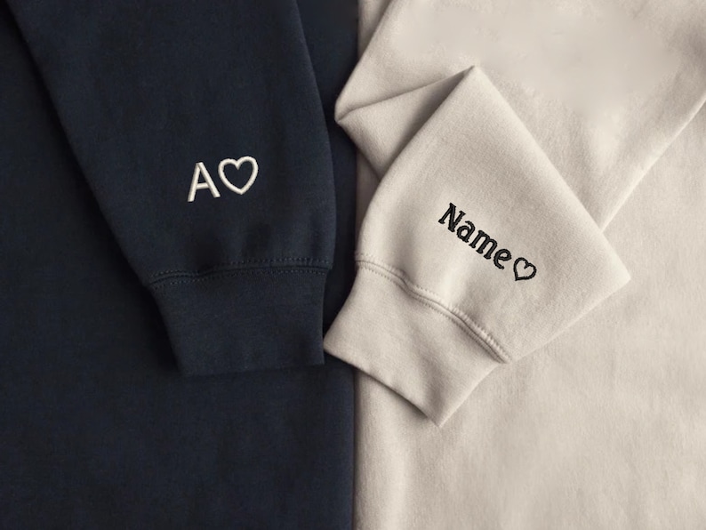 Puede incluir: Dos sudaderas con corazones y texto bordados. La sudadera de la izquierda es azul marino con bordado blanco que dice "A" con un coraz&oacute;n. La sudadera de la derecha es beige claro con bordado negro que dice "Name" con un coraz&oacute;n.