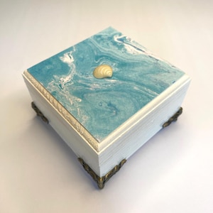 May include: A square wooden box with a white painted base and ornate metal feet. The lid features a blue and white marbled pattern resembling ocean waves, topped with a single small, light-colored seashell centered on the surface.