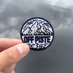 Peut inclure: Un écusson brodé circulaire présentant un graphique de chaîne de montagnes en noir et blanc au-dessus du texte OFF PISTE. Le motif inclut un petit symbole TM sur le côté droit, avec une bordure noire entourant le fond blanc et les broderies noires.