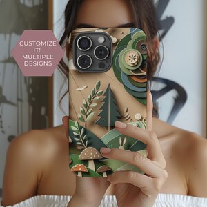 May include: A phone case with a colorful, layered paper design featuring a forest scene with trees, mushrooms, and a rabbit. The case has a cutout for the phone's camera lens.