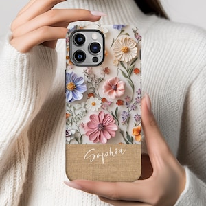 May include: A phone case with a floral design featuring pink, white, and blue flowers. The case has a burlap texture and the name "Sophia" is printed on the bottom.