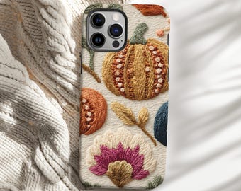 May include: A phone case featuring an embroidered design of a pumpkin, flowers, and leaves in shades of orange, yellow, pink, and blue. The case has a cream-colored background and a camera cutout.