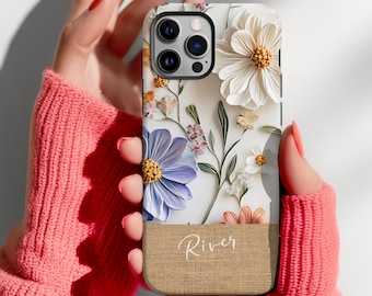 May include: A white iPhone case with a floral design featuring white, blue, and brown flowers. The case has a burlap-like bottom with the word "River" written in cursive.