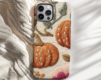 May include: A phone case featuring an embroidered design of two orange pumpkins with white dots, green leaves, and pink and purple flowers. The case has a cream-colored background and a camera cutout.
