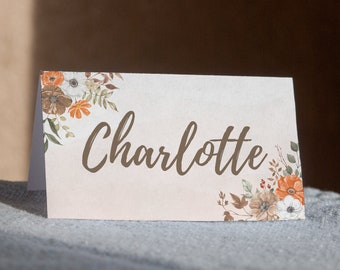 May include: A white place card with a floral design in shades of orange, brown, and white. The name "Charlotte" is written in brown cursive script.