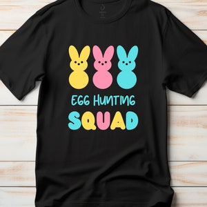 Puede incluir: Camiseta negra con un gráfico de tres siluetas de conejos de color pastel y el texto "Egg Hunting Squad".