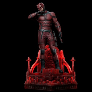 Op de afbeelding: Een rood en zwart Daredevil-beeld staat op een rode basis in gotische stijl tegen een zwarte achtergrond. De figuur staat in een vechthouding, gekleed in een getextureerd pak, laarzen en een masker met hoorns. Het beeld houdt een wapen vast.