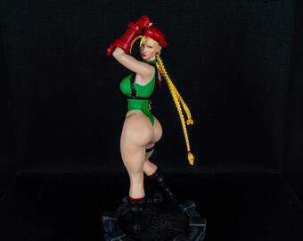 May include: A green and red action figure of a female character with blonde hair in a ponytail. The figure is posed with one arm raised above her head and the other arm bent at the elbow. The figure is standing on a black base with a stone-like texture.
