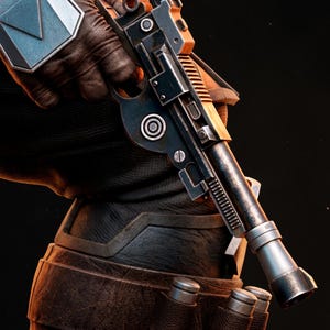 May include: A close-up of a black and silver blaster pistol with orange accents, held by a person wearing a black glove and armor. The pistol has a cylindrical barrel and detailed mechanical components. The person is also wearing brown leather armor.