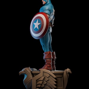 May include: A detailed statue of Captain America in a blue and red costume, holding his iconic shield. The shield is red, white, and blue with a white star. The statue stands on a decorative base with eagle-like carvings, set against a black background.