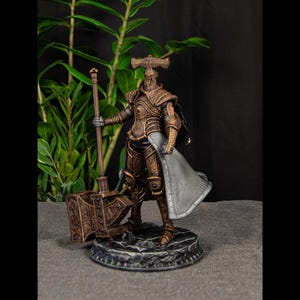May include: A detailed bronze-colored figurine of a knight in full armor, holding a large hammer. The figure stands on a textured base, with a cape draped over one shoulder. The background features green foliage against a dark backdrop.