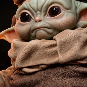 May include: A detailed sculpture of a character with large, expressive eyes and wrinkled skin. The figure is primarily green with large, dark eyes and a slightly furrowed brow. It is wrapped in a tan fabric.