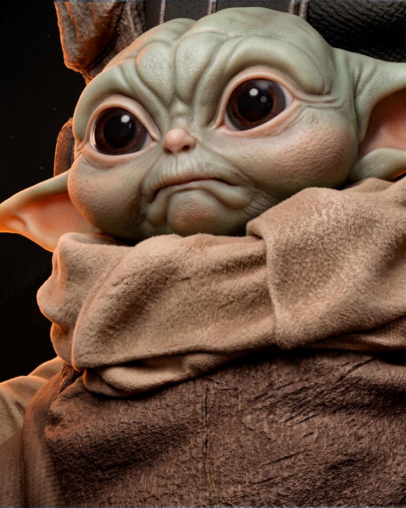 May include: A detailed sculpture of a character with large, expressive eyes and wrinkled skin. The figure is primarily green with large, dark eyes and a slightly furrowed brow. It is wrapped in a tan fabric.