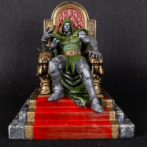 Op de afbeelding: Een zilveren en groene action figure van Doctor Doom die op een gouden troon zit met een rood tapijt. De troon heeft een rode en gouden rugleuning.
