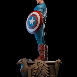 May include: A detailed statue of Captain America in a dynamic pose, holding his iconic shield. The figure is clad in a blue and white costume with red boots. The shield features a white star on a blue field, surrounded by red and white rings. The statue rests on a decorative base.