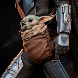 May include: A detailed image of a green-skinned, large-eyed creature nestled in a brown pouch. The creature is held by a person wearing dark armor with silver accents. The scene is set against a dark background, highlighting the details of the character and armor.