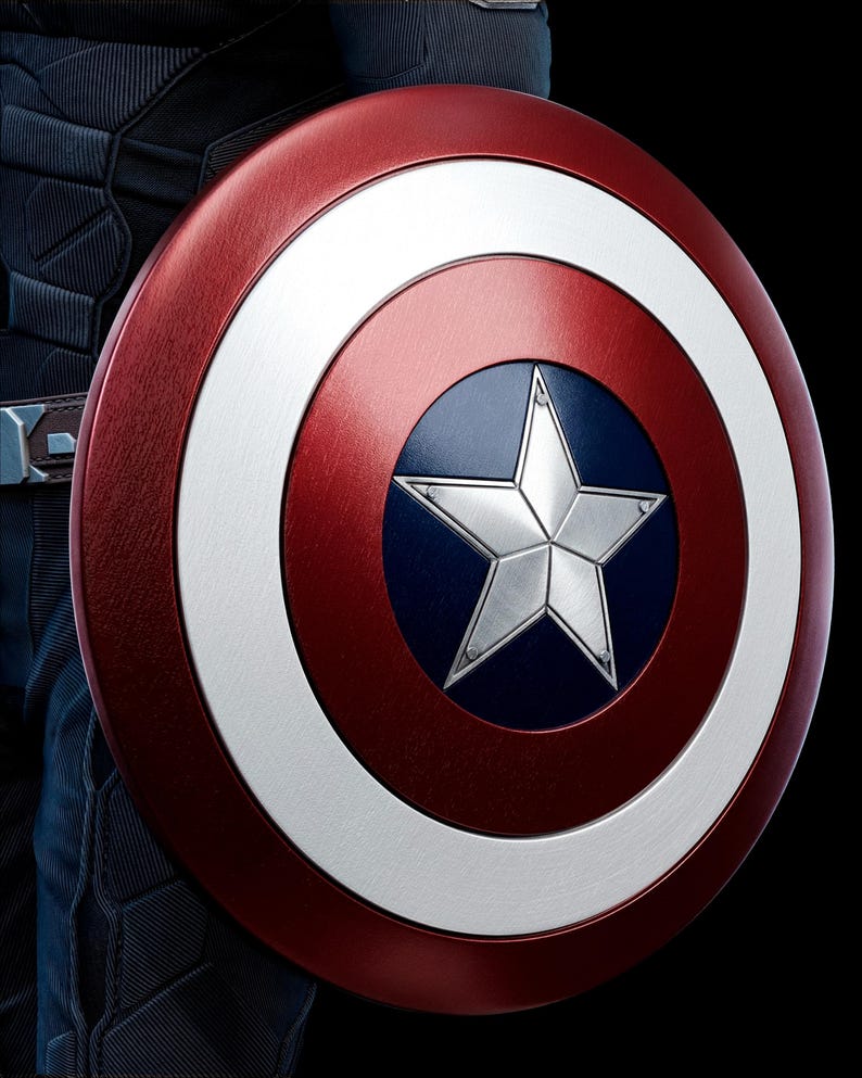 May include: A close-up of Captain America's shield. The shield is circular, with a red outer ring, a white inner ring, and a blue center. A silver star is in the center. The shield is held against a dark background.