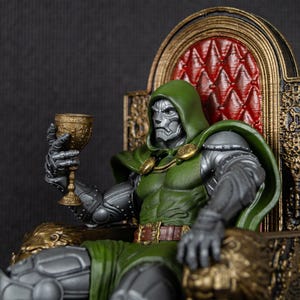 May include: A green and silver action figure of Doctor Doom sitting on a gold throne, holding a silver goblet. The figure is wearing a green cape and a silver mask.