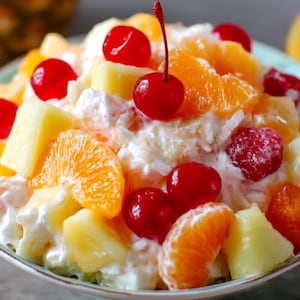 May include: A vibrant fruit salad in a light green bowl. The salad features pieces of pineapple, orange segments, and red cherries. The fruit is mixed with a creamy white dressing and garnished with shredded coconut.