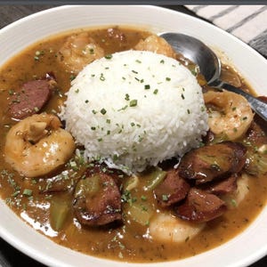 May include: A white bowl filled with a brown stew with shrimp, sausage, and green vegetables. A mound of white rice is on top of the stew.