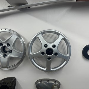 Può includere: Tre cerchi in lega d'argento per auto con cinque razze ciascuno sono montati su una parete bianca. Il centro del cerchio a destra ha un logo "OZ" nero. Un mozzo in metallo nero è montato sulla parete a destra dei cerchi.