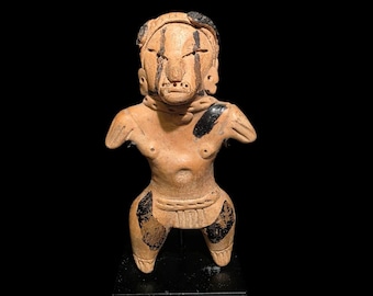 May include: A terracotta figurine of a standing figure with black painted details. The figure has a stylized face with black markings and is adorned with black accents on the shoulders and legs. The figurine is displayed on a black stand against a black background.