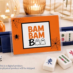 Pode incluir: Um cartão retangular laranja numa capa de plástico transparente com ilustrações de teias de aranha e aranhas pretas, com o texto BAM BAM BOO em laranja e preto, onde os dois Os são substituídos por ícones de fantasmas brancos.