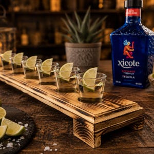 Puede incluir: Una tabla de servir rectangular de madera con acabado carbonizado, que presenta seis vasos de chupito transparentes llenos de tequila y decorados con rodajas de lima. Una botella de cristal azul de tequila Xicote se encuentra al lado.