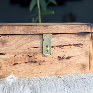 May include: A wooden box with a rectangular shape and a hinged lid. The box is made of light-colored wood with a natural grain pattern and dark brown accents. A brass-colored latch secures the lid. The box is sitting on a white surface.