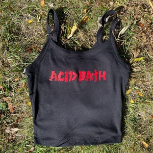 Puede incluir: Camiseta sin mangas negra con tirantes finos. Las palabras "ACID BATH" están impresas en rojo en la parte delantera. La camiseta está sobre hierba verde.