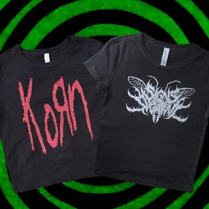 Puede incluir: Dos camisetas negras. Una presenta la palabra "Korn" en letras rojas desgastadas. La otra muestra un diseño de insecto blanco estilizado con el nombre de la banda. Ambas camisetas tienen mangas cortas y cuello redondo.