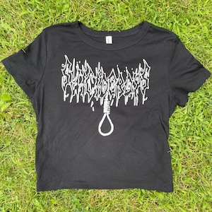 May include: Black crop top with white graphic text and a noose design. The text is stylized and appears above the noose. The shirt is made of a soft material and is displayed on a bed of green grass.