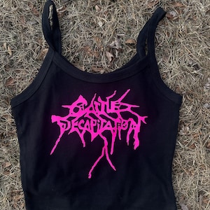 Puede incluir: Camiseta sin mangas negra con las palabras "Cattle Decapitation" en letras rosas brillantes y estilizadas. La camiseta tiene tirantes finos y un escote redondo. La imagen está tomada al aire libre sobre hierba seca.