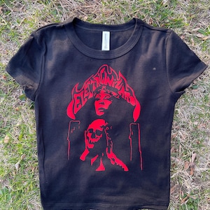 Könnte beinhalten: Schwarzes Crop-Top mit rotem Grafikdesign. Das Design zeigt den Text "ELECTRIC WARRIORS" über dem Gesicht einer Frau und einem Totenkopf, mit Flammen und Kerzen. Das Shirt ist aus weichem Material.