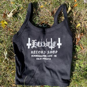 May include: Black tank top with white text that reads "Helvete Record Shop, Schweigaards Gate 56, Oslo Norway!!" The top has a scoop neck and thin straps. The top is laying on a bed of green grass.