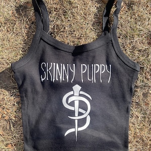 Peut inclure: Débardeur noir à fines bretelles. Le haut présente le texte blanc "SKINNY PUPPY" dans une police gothique au-dessus d'un graphisme blanc d'une lettre "S" stylisée avec une épée qui la traverse. Le haut est fait d'une matière douce.