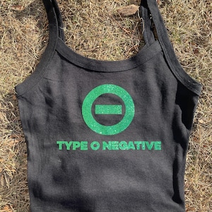 以下が含まれることがあります： 緑色のグリッターロゴと「TYPE O NEGATIVE」の文字が入った黒のクロップトップ。細いストラップとラウンドネックラインが特徴です。デザインは前面の中央に配置されています。