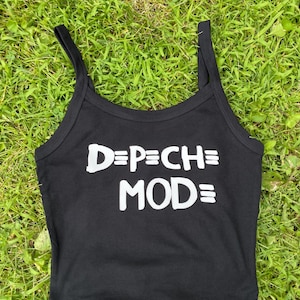 May include: Black tank top with the text "DEPECHE MODE" in white. The top has thin straps and a scoop neckline. The tank top is made of a soft material and is suitable for casual wear.