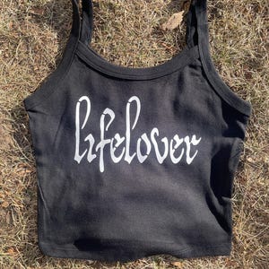 May include: Black crop top with thin straps and the white text "lifelover" in a cursive font. The top is made of a soft material and is suitable for casual wear. The garment is displayed on a bed of dry grass.