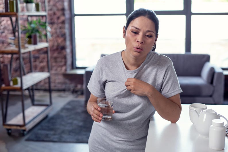 Może przedstawiać: Osoba w szarej koszulce trzyma szklankę wody, wyglądając na chorą. Biały czajnik, filiżanka i butelki z pigułkami stoją na białym blacie. Metalowa p&oacute;łka z roślinami i szary dywan są w tle.