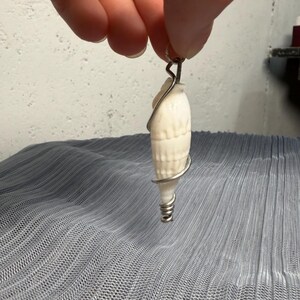 以下が含まれることがあります： 銀色の金属ワイヤーで巻かれた、白く細長い螺旋状の貝殻ペンダント。ワイヤーは上部で小さなループを形成して吊り下げられるようになっており、貝殻の尖った底部の周りにしっかりと巻き付いて固定されています。