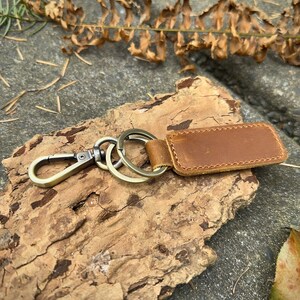 May include: A brown leather keychain with a rectangular tag and metal hardware. The keychain features a clasp and multiple rings. The leather tag is stitched and has a natural, textured appearance. The hardware has a bronze finish.
