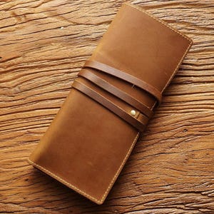 May include: A brown leather wallet with a strap closure. The wallet is rectangular and has a textured surface. The strap is wrapped around the wallet and secured with a brass-colored stud. The wallet is on a wooden surface.
