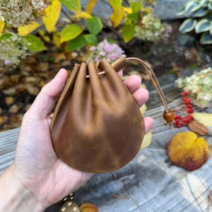 May include: A brown leather drawstring pouch, held in a hand, with a leather cord and wooden bead closure. The pouch is round and gathered at the top. The background includes fall foliage and a gold-colored die.